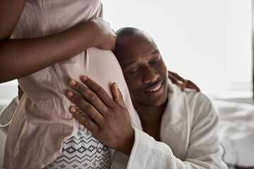 happy young african man is listening to the belly of his pregnant wife, couple in domestic wear at home