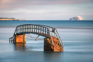 The Belhaven Bridge, Dunbar, Scotland.. Also known as the Bridge to Nowhere, as although it crosses the River Biel, its base is submerged at high tide. The Bass Rock can be seen in the distance. - obrazy, fototapety, plakaty