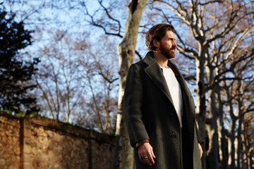 Fototapeta na wymiar Portrait of fashionable well dressed man with beard posing outdoors looking away, confident and focused mature man in coat standing outside at sunny evening, elegant fashion model