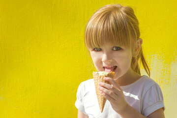 Little cute girl with emotion on her face eating an ice cream cone on the background of a yellow bright wall, copy space for text