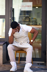 upset african man sit in the street, his cafe shop is crashing, it is closed, in crisis. black man...