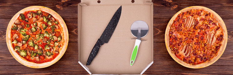 In an open cardboard pizza box is a black kitchen knife and a pizza cutter, next to it are two pizzas with meat and vegetables, on the wooden rustic table, top view
