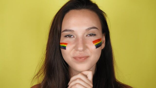 A woman with a rainbow pattern on her face is looking at the camera. The LGBT flag is painted on the face. Yellow background.