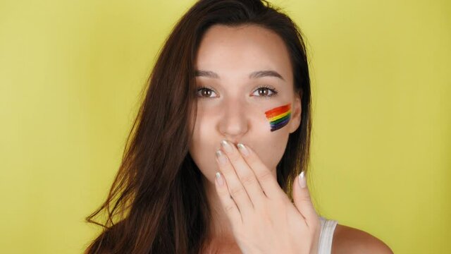 Happy woman with a rainbow pattern on her face sends an air kiss. The LGBT flag is painted on the face. Yellow background.