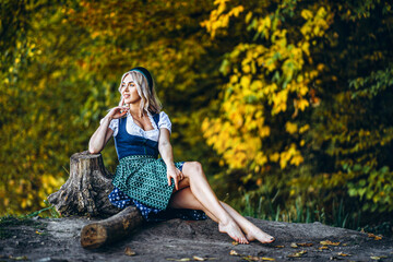 Barefoot happy pretty blond girl in dirndl, traditional beer festival dress, sitting outdoors with blured colorful trees behind