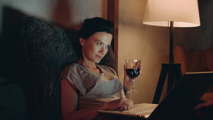 Girl surfing internet on notebook on couch. Woman drinking red wine with laptop.