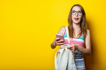 Beautiful young woman in glasses with a linen bag holding a phone on a yellow background.