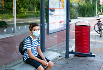 a caucasian schoolboy sitting on the bus stop waiting for a bus wearing a protective face mask. Protection during quarantine