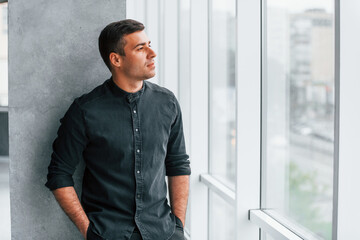 Elegant man in black shirt leaning on the wall indoors and looking through the big window