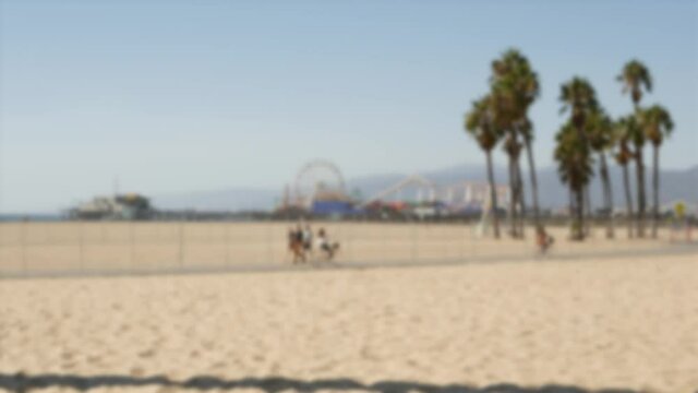 California beach aesthetic, people ride cycles on a bicycle path. Blurred, defocused background. Amusement park on pier and palms in Santa Monica american pacific ocean resort, Los Angeles CA USA.