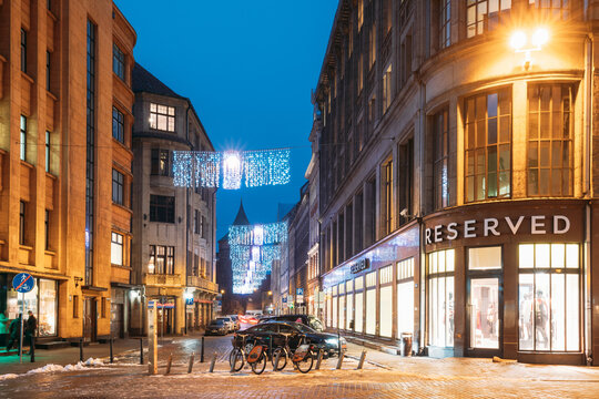 Riga, Latvia. Row Of Bicycles For Rent At Bike Parking Near Clothing Shop The Reserved On Valnu Street In Lighting In Night Illumination. Reserved Is A Polish Clothing Store Chain