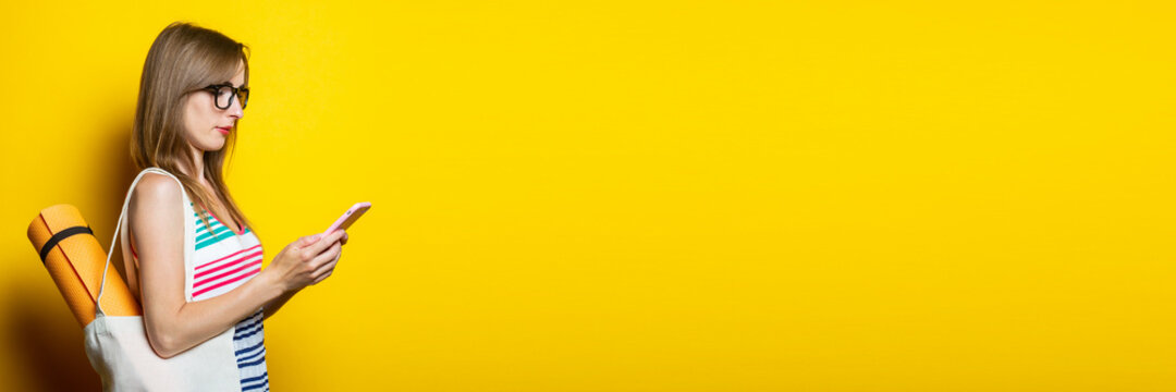 Beautiful young girl with karimat in a bag looks at the phone on a yellow background. Banner.