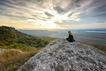 A woman on the top of a rock enjoys the view of sunset and using digital tablet