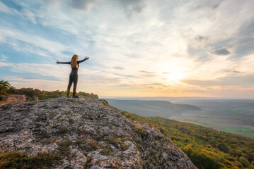 A woman on the top of a rock enjoys the view of sunset over an autumn forest