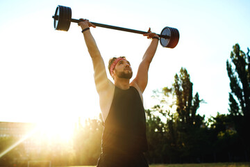 Man athlete lifts the barbell over his head, training athlete outdoors at sunset