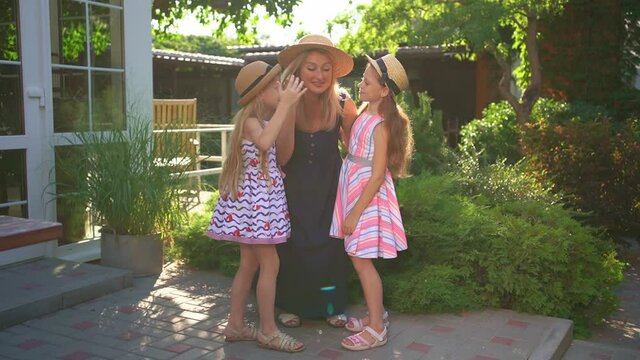mother with daughters in hats embrace and kiss in garden at backyard of house