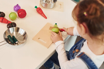 Little girl in casual clothes sits by table and having fun by playing with toys on the kitchen