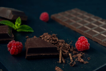 Pieces of chocolate, chocolate shavings, raspberry and mint on dark textured background.