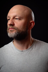 Studio portrait of a bald man with beard in his 40s. Model dressed in simple grey t shirt looking away from the camera. Black background.