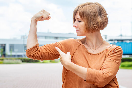 Adult redhead woman in casual clothes standing demonstrates muscles flexing her arm. The concept is a strong and independent modern woman.
