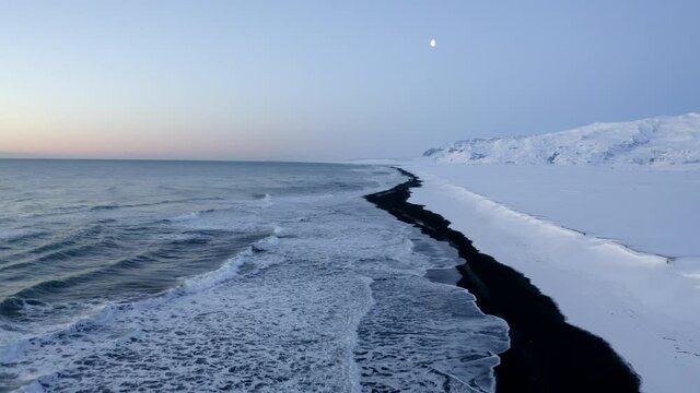 AERIAL: Flying over Black Beach with white arctic snow mountains in background in Iceland in Winter Snow, Ice, Waves, Water