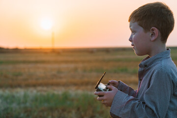 Red-haired boy at sunset in the evening stands on a wheat field with bales and controls the drone
