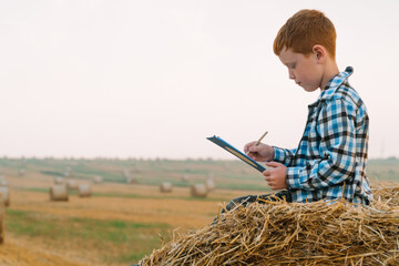 Naklejka na ściany i meble A red-haired boy on top of a straw bale holds a tablet with a letter and a pen in his hands on a background of hay bales