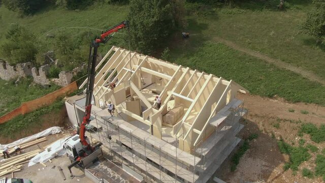 AERIAL: Flying around a modern prefabricated house being built in the Slovenian countryside. Group of contractors mounts a wooden arch on the roof of a cross-laminated timber house under construction.