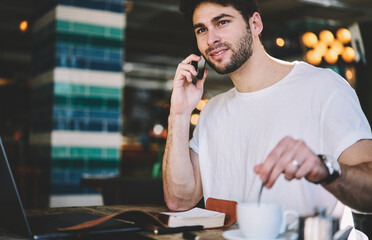 Good looking freelancer nicely talking on the mobile phone with cup of coffee while studying on modern laptop with 5G internet connection using notebook and pen for records, man looking out of window