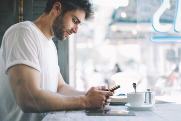 Cropped image bearded hipster guy check own mail and chatting with friends in social networks on mobile phone using free internet sitting in cafe shop with cup of coffee.  Advertising copy space area