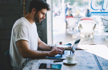 Caucasian young skilled concentrated student typing text on modern laptop and searching new ideas for coursepaper using digital devices and 5G wireless internet sitting in urban cafeteria during break