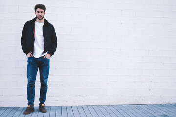 Full length portrait of stylish hipster guy standing on white promotional background for your advertising text message.Handsome man dressed in casual outfit looking at camera near copy space area