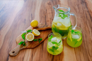Vintage jug with two glasses of refreshing non alcoholic mojito lemonade drink with lemon slices, mint leaves, straw & ice on wooden kitchen table. Close up, copy space, background