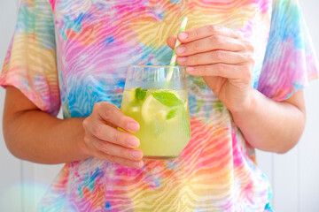 Young woman wearing a tie dye t-shirt holding a glass of iced lemonade. Female with refreshing non alcoholic mojito drink with lemon slices, mint leaves & ice on white background, close up, copy space