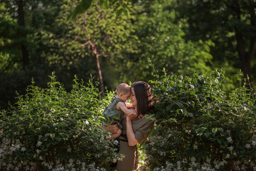 Naklejka na ściany i meble A young woman holds a little baby in her arms. A beautiful mother walks with her daughter in a green park near the jasmine bushes. Mother's day, motherhood concept, mother love is limitless. Close up
