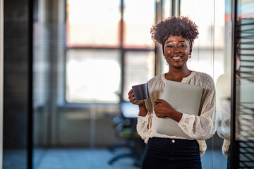 Beautiful young grinning professional Black woman in office with eyeglasses, folded arms and confident expression. Smiling smart attractive young black lady standing in lobby and looking at camera