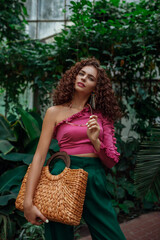 Fototapeta na wymiar Fashionable curly woman wearing pink asymmetric ruffled top, green trousers, holding straw wicker top handle bag, posing in tropical garden. Summer fashion, lifestyle, beauty conception