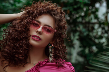 Curly brunette woman with fuchsia color lips makeup, afro hairstyle, wearing pink glasses, big earrings, posing in tropical garden. Summer fashion, beauty conception. Copy, empty space for text