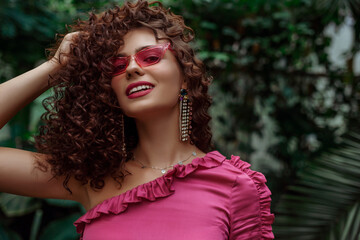 Happy smiling curly woman with fuchsia color lips makeup, wearing pink glasses, big earrings,...