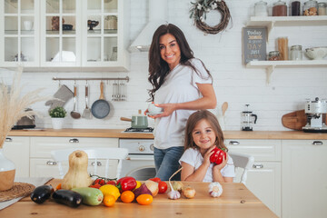 Happy family mother and child posing at home. Beautiful young mom and little daughter having fun and preparing vegetables for salad in a white kitchen in a Scandinavian style interior. Healthy food.