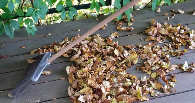 Cleaning fallen dry autumn leaves on a wooden floor using a large broom with plastic bristles.