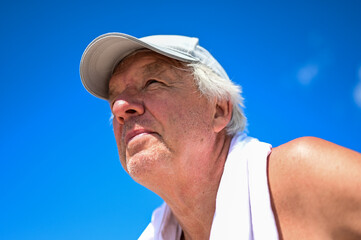 Close up of a retired man with gray hair wearing a cap and a towel around his neck, looking into the distance.