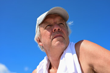 Close up of a senior man with gray hair wearing a cap and a towel around his neck, looking into the distance.