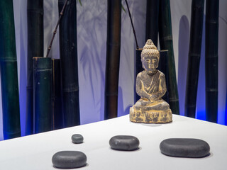 Buddha figure in a spa with massage stones and relax ambiance. Buddha sculpture sit in Lotus position in a beauty salon with bamboo in the background. Spirituality and zen concept