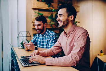 Cheerful male student laughing at funny joke learning on laptop computer spending time in campus with friend,happy prosperous freelancers satisfied with productive job using modern technologies