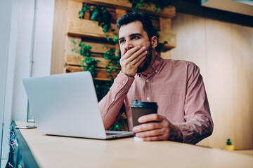 Fototapeta na wymiar Tired male freelancer sitting near laptop computer yawning feeling exhausted with completing job before deadline, bored hipster guy tired with hard exam preparation leaning with netbook and coffee