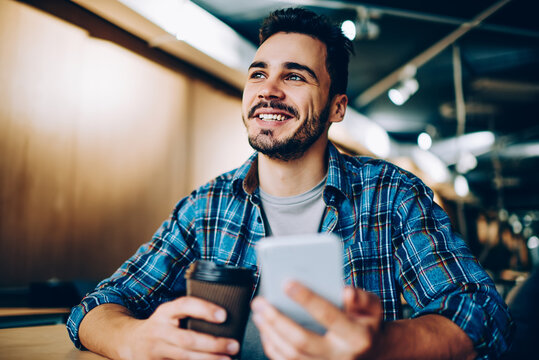 Cheerful Handsome Guy Looking Up Imagine His Vacation While Chatting Via Smartphone In Office, Young Smiling Student Dreaming About Something On Coffee Break Holding Mobile Phone For Networking