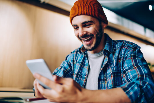 Overjoyed male in hat surprised with victory in online competition in social networks getting notification on cellular, amazed hipster guy happy about winning discount for online courses studying .