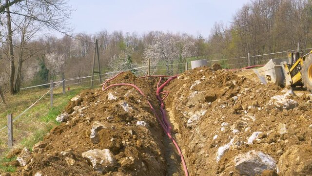 CLOSE UP: Corrugated plastic telecommunication tubing runs along a deep trench and up the green hill near a busy construction site. Wide red conduits supply the scenic countryside with electricity.