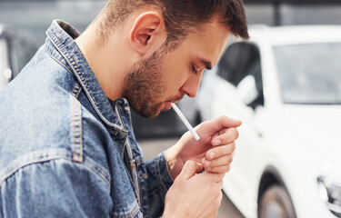 Fototapeta premium Fashionable man smoking near car outdoors against modern business building